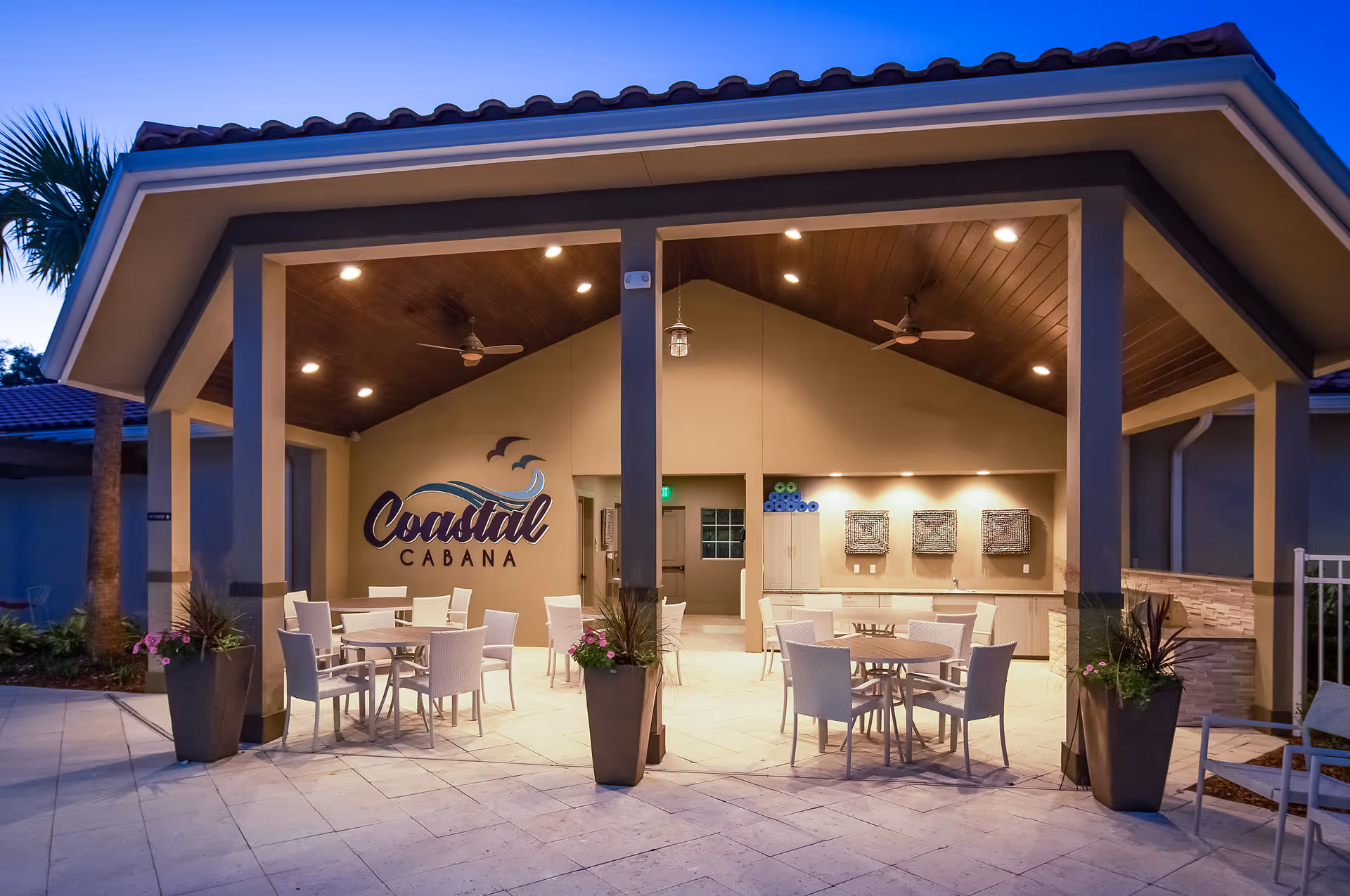 Outdoor covered seating area at dusk with round tables and white chairs under a wooden ceiling with ceiling fans. The wall features a sign that reads 'Coastal Cabana' with a wave and birds graphic. There are potted plants on either side of the entrance and rolled towels stacked on a cabinet in the back.