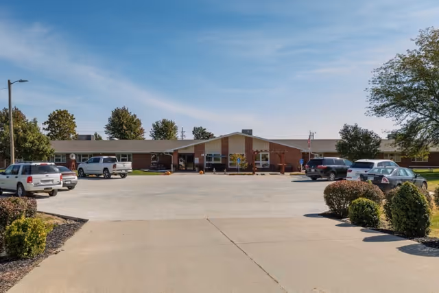 Front exterior view of Falls City Nursing and Rehabilitation Center building with a parking lot in front, several parked cars, and trees surrounding the area under a clear blue sky.