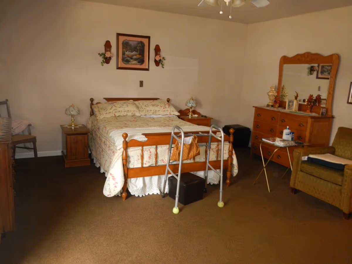 A senior living bedroom with a wooden bed featuring floral bedding, two matching nightstands with lamps, a wooden dresser with a large mirror, an armchair, a small side table with hand sanitizer and tissues, and a walker positioned at the foot of the bed.
