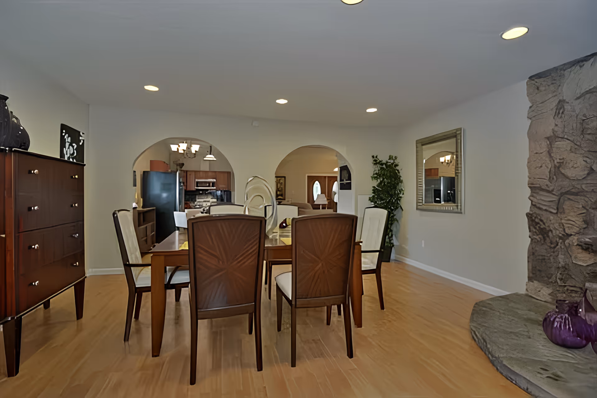 Dining room with a wooden table and chairs, arched openings to the kitchen, a stone fireplace on the right and hardwood floors.