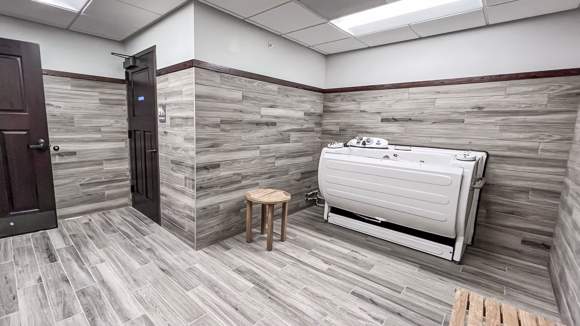 A room with wood-patterned tile flooring and walls partially covered with matching wood-patterned tiles. There is a white walk-in bathtub with control knobs on the right side of the room, a small wooden stool next to it, and two dark wooden doors on the left side. One door has a sign indicating a restroom.