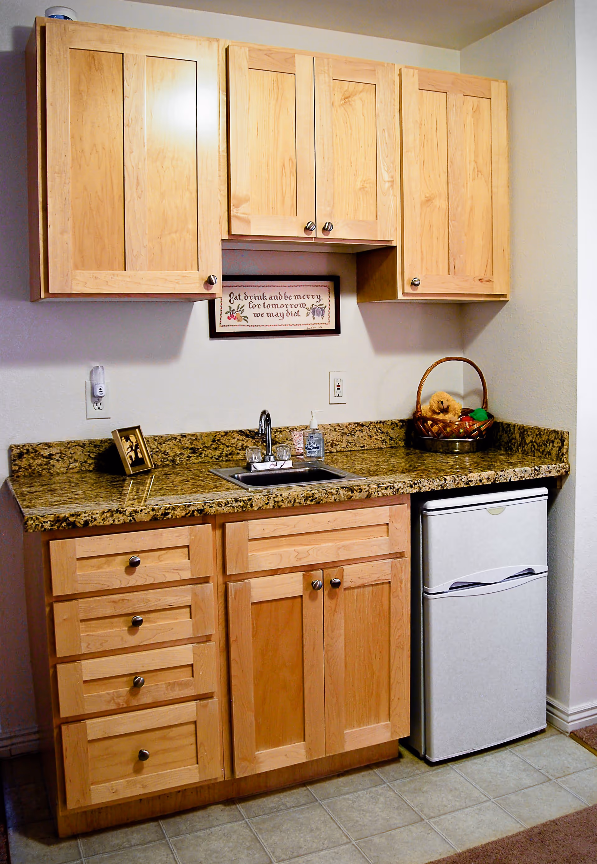 A small kitchenette with light wood cabinets, a granite countertop, a sink, and a compact refrigerator.