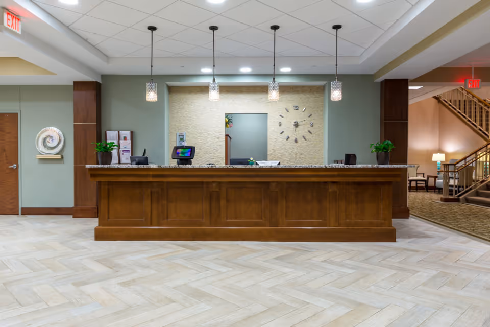 Reception desk area in a senior living facility with a wooden counter, three pendant lights hanging from the ceiling, a wall clock, potted plants, and a seating area with stairs visible in the background.