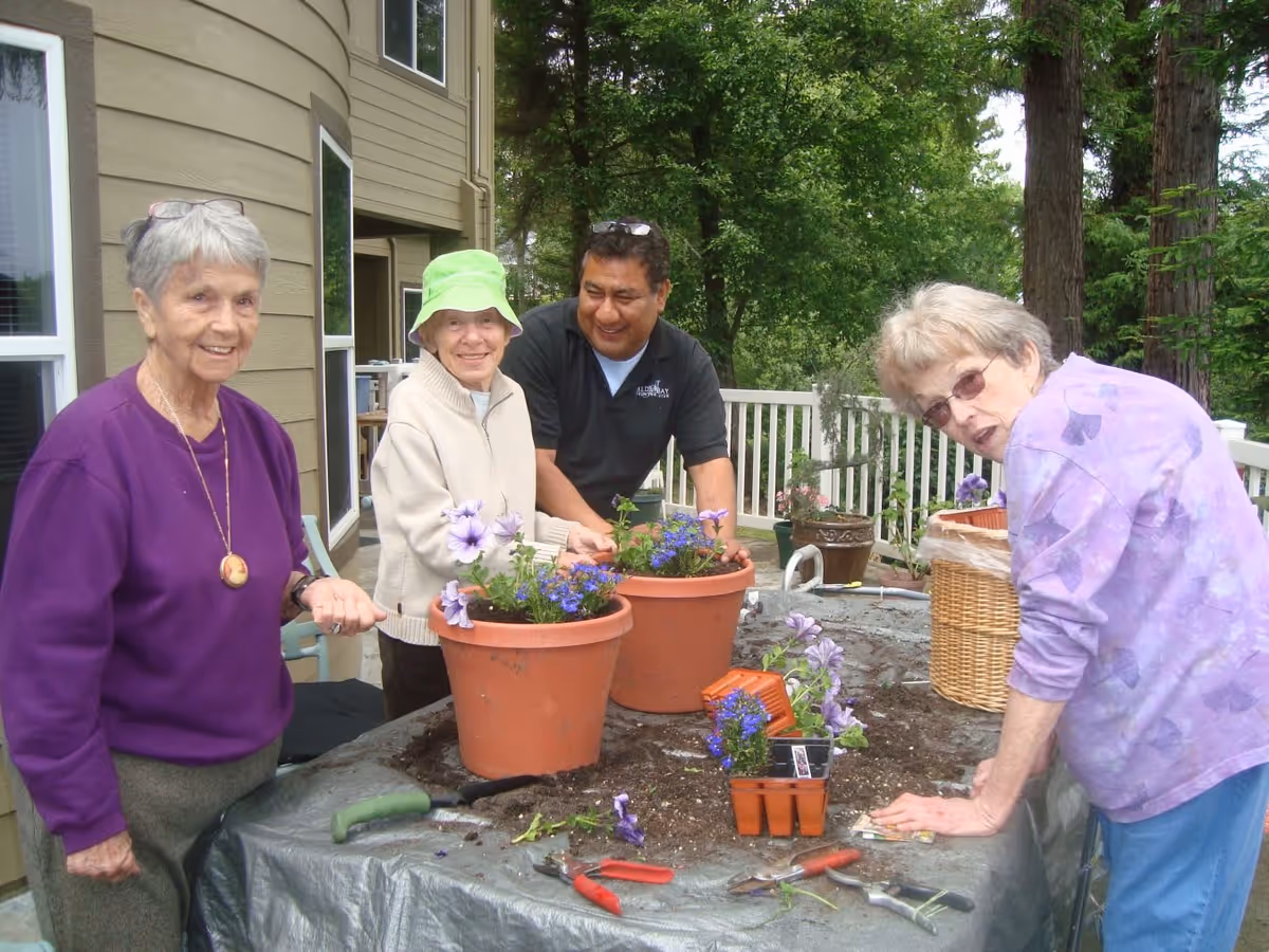 Three elderly women and one man are gardening together on a patio. They are planting purple flowers in large terracotta pots on a table covered with a protective sheet. The background shows a building and lush green trees.