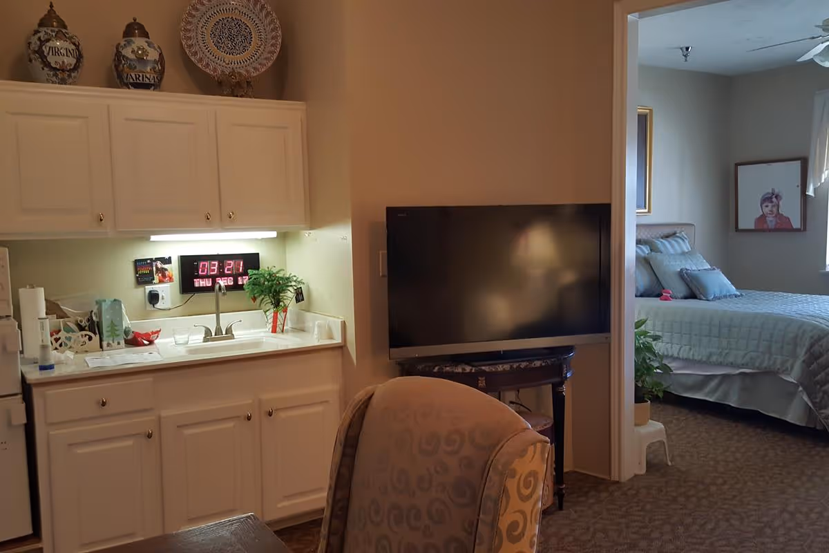 Interior view of a senior living facility room showing a small kitchenette with white cabinets, a sink, and decorative items on top. A digital clock displaying the time and date is mounted under the cabinets. To the right, there is a flat-screen TV on a small table and an armchair with a patterned fabric. In the background, a bedroom is visible with a neatly made bed, pillows, a framed picture on the wall, and a window with curtains.