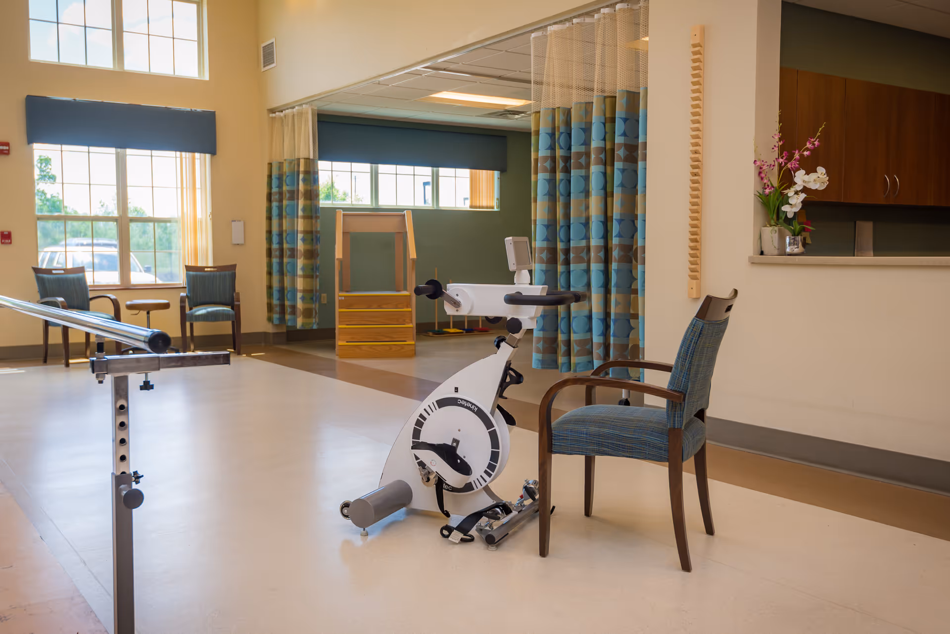 A bright rehabilitation room with a stationary exercise bike, a chair, parallel bars and seating by large windows.