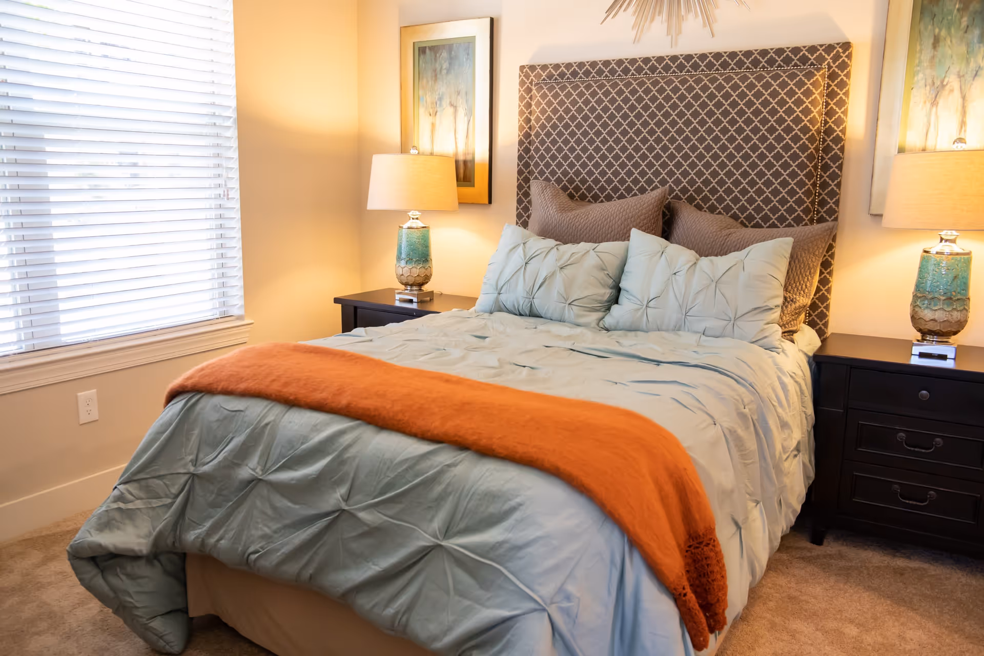 A neatly made bed with a patterned headboard, light blue bedding, and an orange throw blanket at the foot. There are two dark wooden nightstands on either side of the bed, each with a lamp and a framed painting above. A window with white blinds is on the left side of the room.