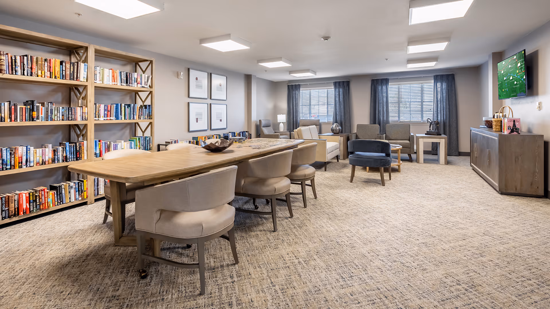 Communal lounge and library with a long wooden table surrounded by chairs, bookshelves along the wall, and a seating area with sofas and a TV.