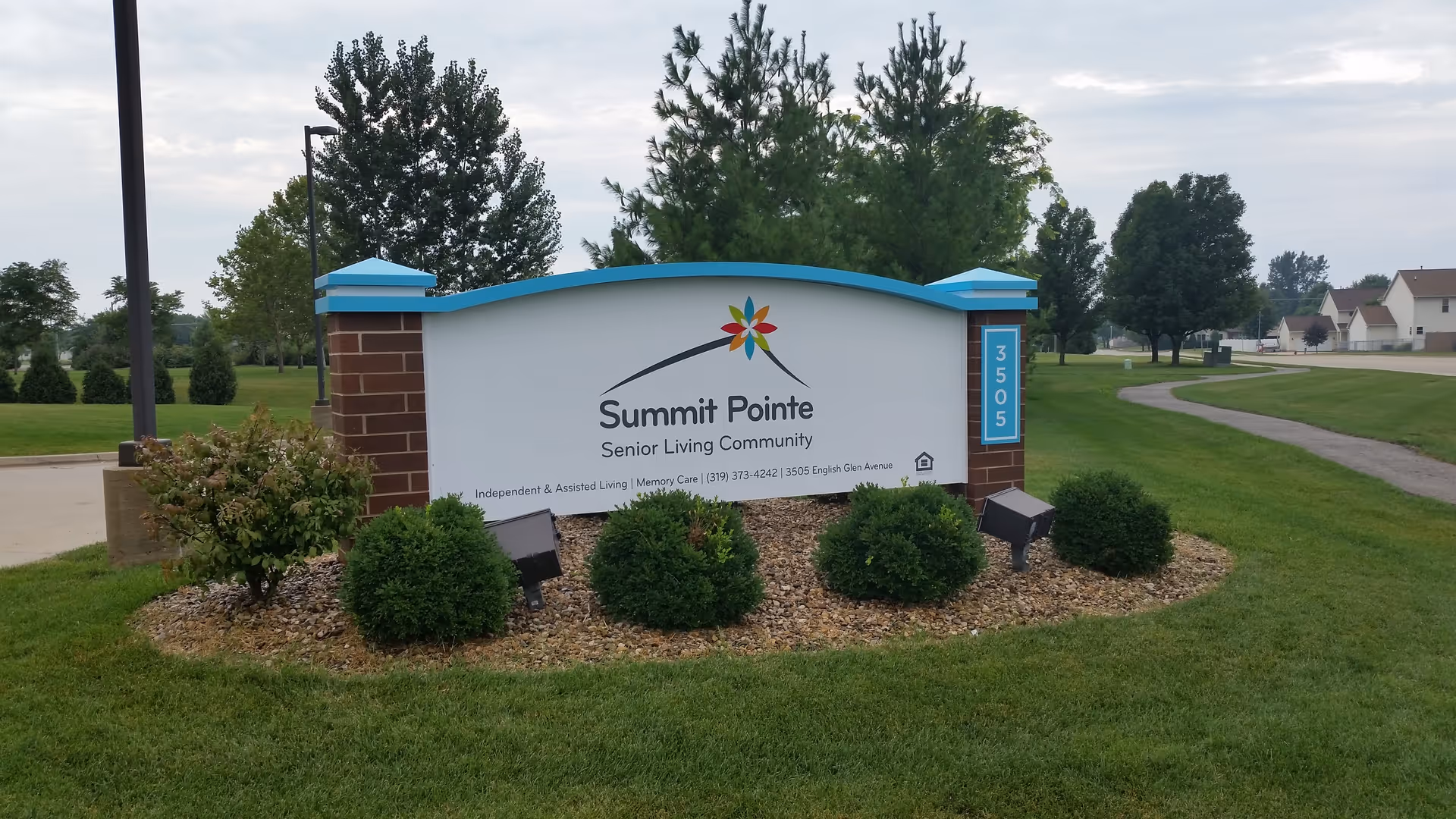 Outdoor image of a Summit Pointe Senior Living Community sign surrounded by green grass, bushes, and trees with a cloudy sky in the background.