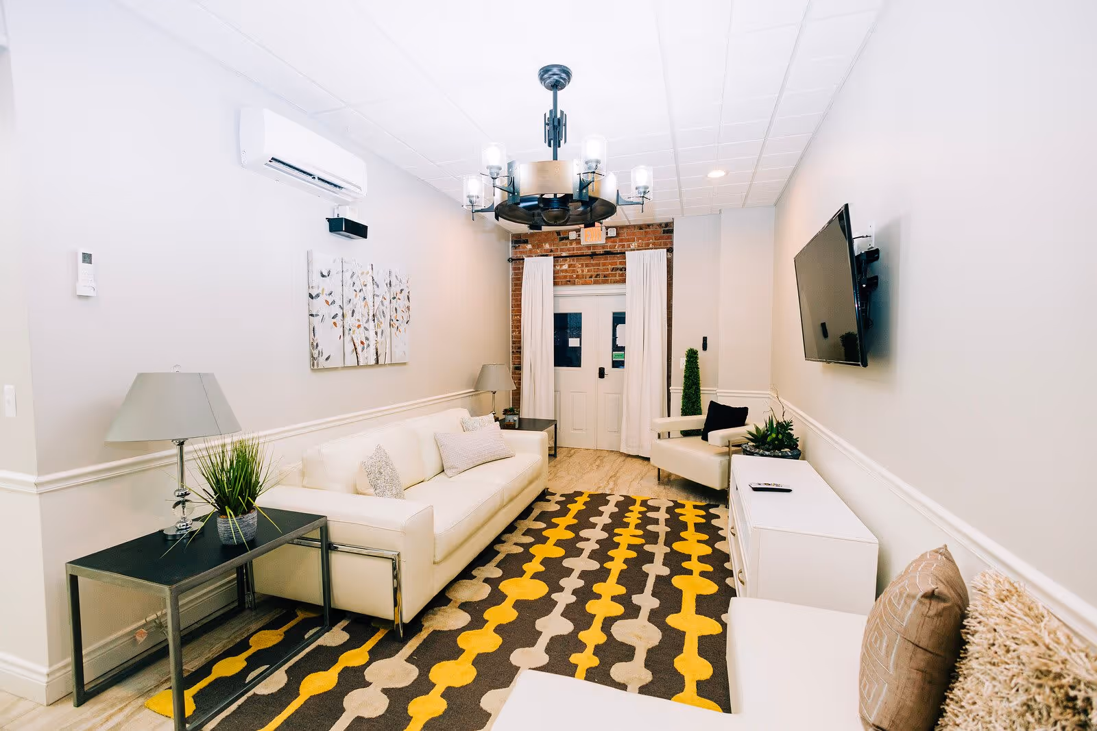 A modern living room with a white sofa, armchair, and a patterned rug in shades of yellow, gray, and beige. The room features a wall-mounted flat screen TV, two side tables with lamps and plants, a chandelier, and a white door with curtains. The walls are light-colored with a brick accent above the door.
