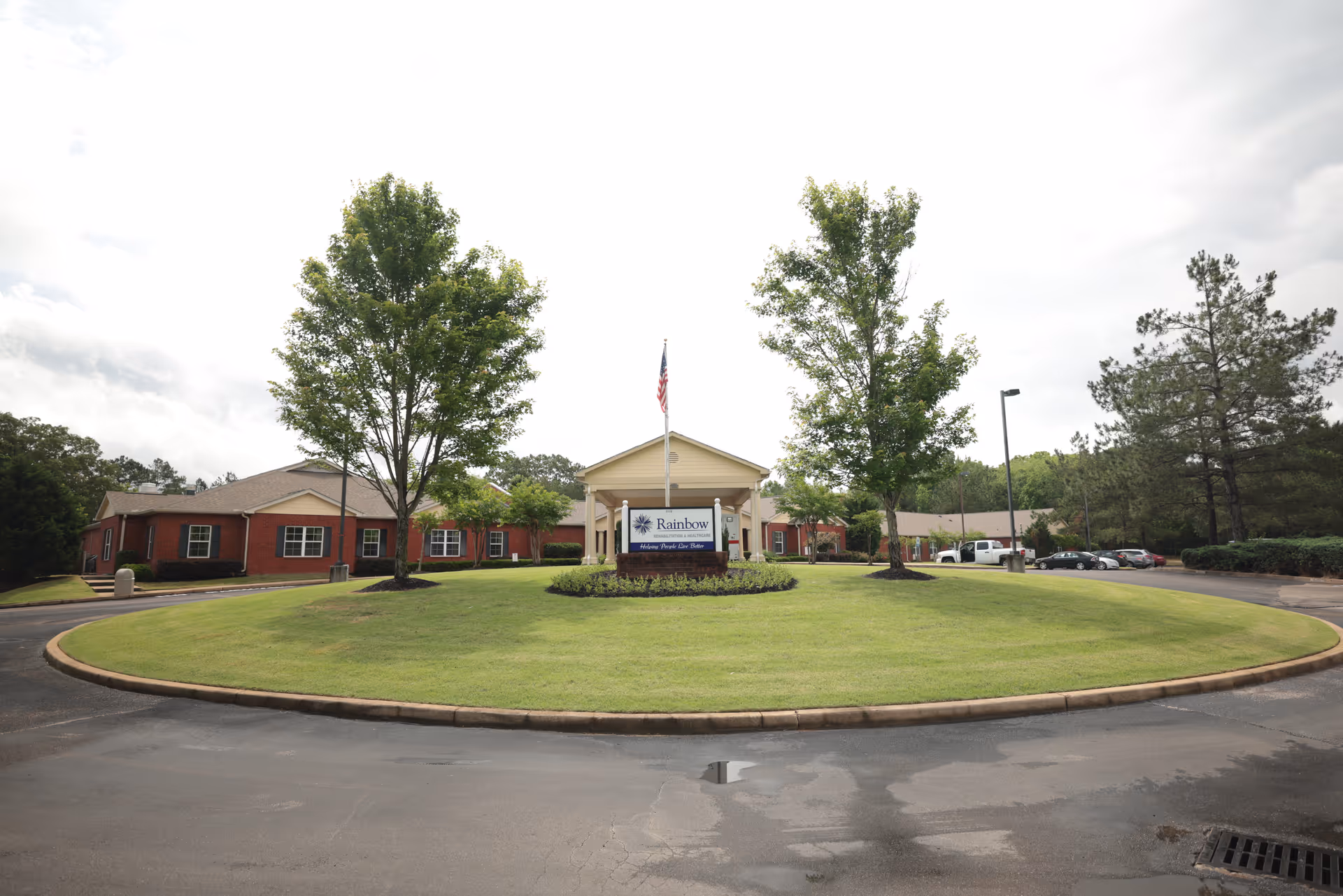Front exterior view of Rainbow Rehab and Healthcare facility with a circular driveway, green lawn, trees, and an American flag on a flagpole in front of the building.
