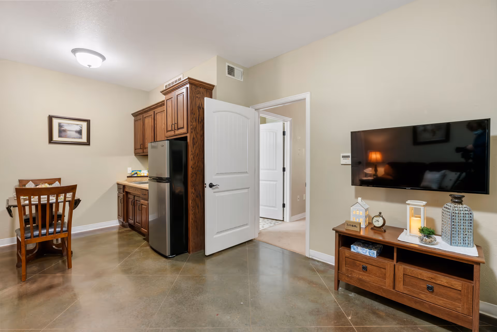 Open living area with a wall-mounted TV above a wooden console, a small dining table, and a kitchenette with dark wood cabinets and a stainless refrigerator.