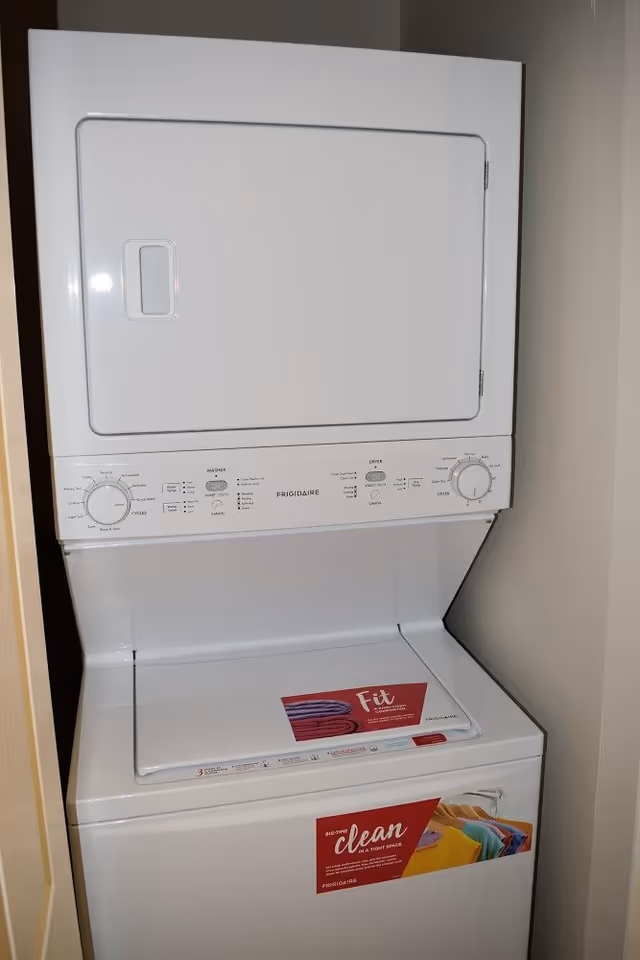 A white stacked washer and dryer unit in a small laundry closet with beige walls. The washer is on the bottom and the dryer is on top. There are informational stickers on the washer lid and front.