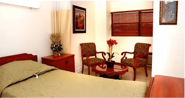 A cozy bedroom in a senior living facility featuring a bed with a green bedspread, a wooden nightstand with a floral arrangement, two patterned armchairs around a small round wooden table with a flower vase, framed artwork on the walls, and a window with wooden blinds.