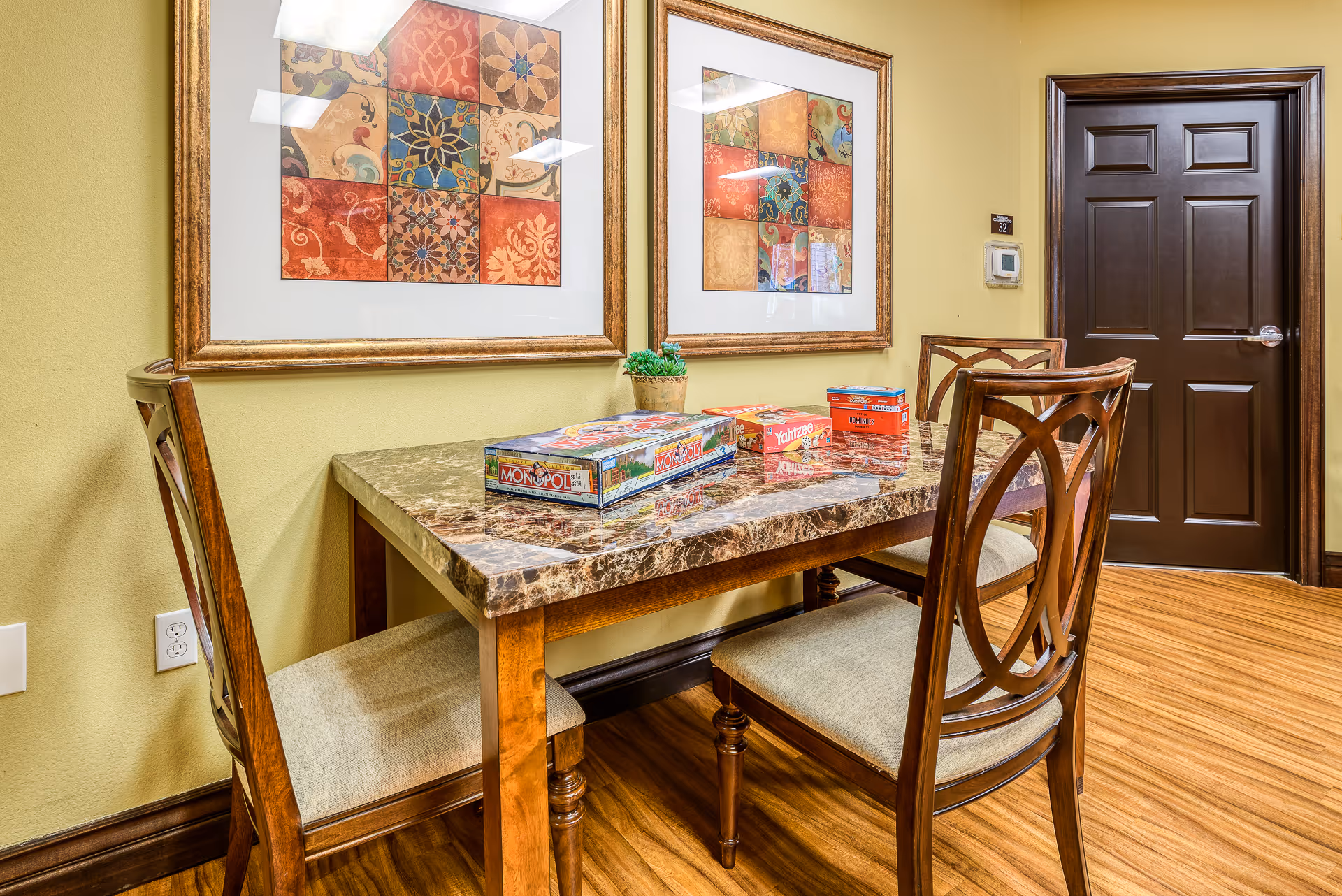 Small dining/game area with a marble-top table, wooden chairs, board games on the table, framed artwork on the wall, and a dark brown door.