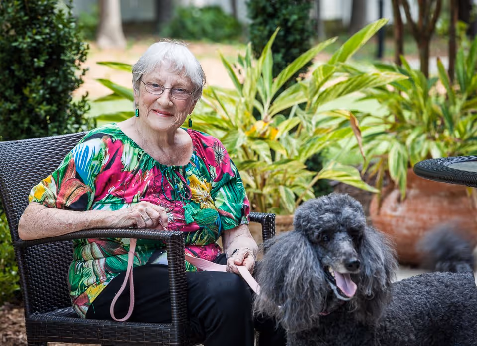 An elderly woman with short white hair and glasses is sitting on a wicker chair outdoors, holding a leash attached to a large gray poodle. She is wearing a colorful floral blouse and smiling. Behind her are green plants and a large terracotta pot.