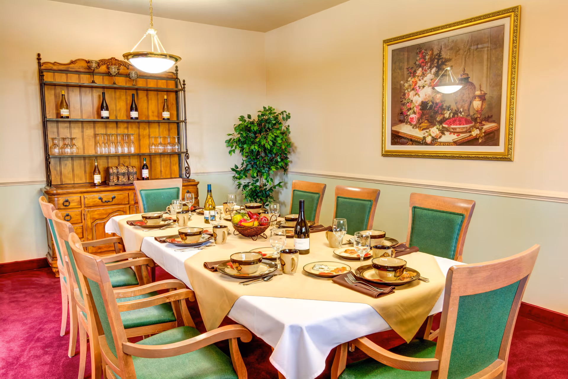A dining room with a rectangular table set for eight people. The table has a white tablecloth with a beige overlay and is set with plates, bowls, mugs, wine glasses, and silverware. There are two bottles of wine on the table and a fruit basket centerpiece. The chairs have wooden frames with green upholstered seats and backs. In the background, there is a wooden hutch with wine bottles and glasses, a potted plant, and a framed floral painting on the wall. The room has a red carpet and cream-colored walls.