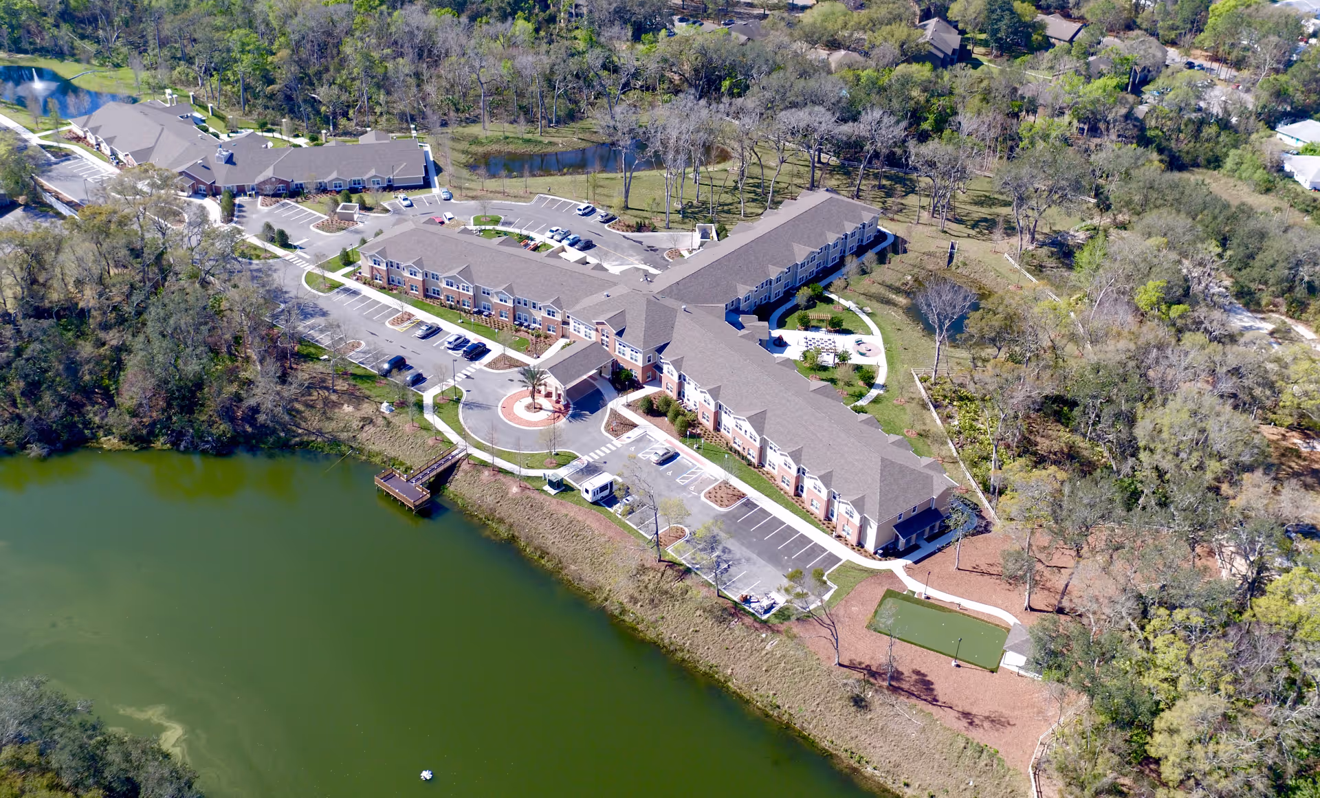 Aerial view of Anthem Lakes senior living facility showing a large, multi-wing building surrounded by trees and greenery, with a parking lot and a small dock extending into a nearby lake.