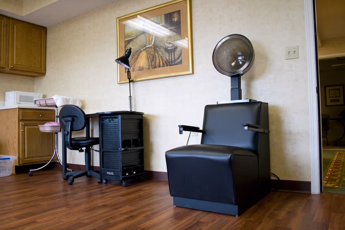 A salon-style interior with a black hair-drying chair under a hood dryer, a small desk and chair, cabinets, and framed artwork.
