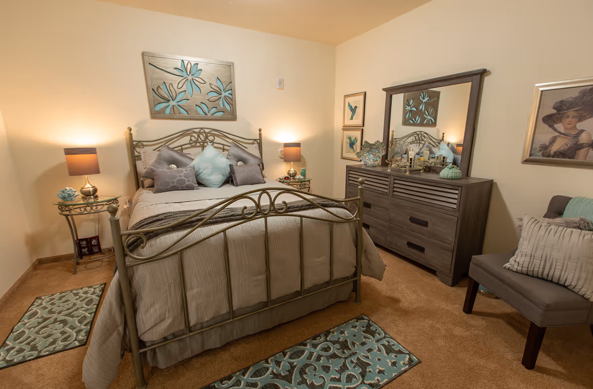 A cozy bedroom with a metal frame bed adorned with multiple pillows in shades of gray and teal. The room features two bedside tables with lamps, a wooden dresser with a large mirror, decorative items, and framed artwork on the walls. There is a cushioned chair with pillows and two patterned rugs on the carpeted floor.