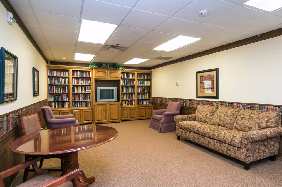 A cozy living room area in an assisted living facility featuring a floral patterned sofa, two purple armchairs, a wooden round table with chairs, and a large wooden bookshelf filled with books and a television in the center. The room has beige walls with decorative wallpaper trim and framed artwork, and a carpeted floor.
