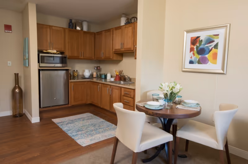 A small kitchen area with wooden cabinets, a microwave, a mini refrigerator, and a countertop with various kitchen items. Adjacent to the kitchen is a round dining table set for three with white chairs and a vase of white flowers. A colorful abstract painting hangs on the wall above the dining table.