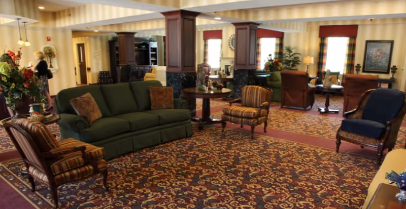 A spacious senior living facility common area with patterned carpet, green sofa, multiple armchairs, wooden tables, and decorative plants. The room has large windows with plaid curtains and warm lighting. A person is seen walking in the background near the hallway.
