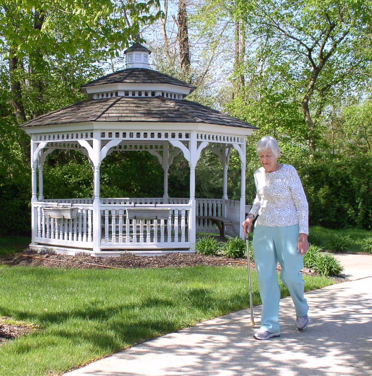 An elderly woman walking with a cane on a paved path next to a white wooden gazebo surrounded by green grass and trees in a garden area.