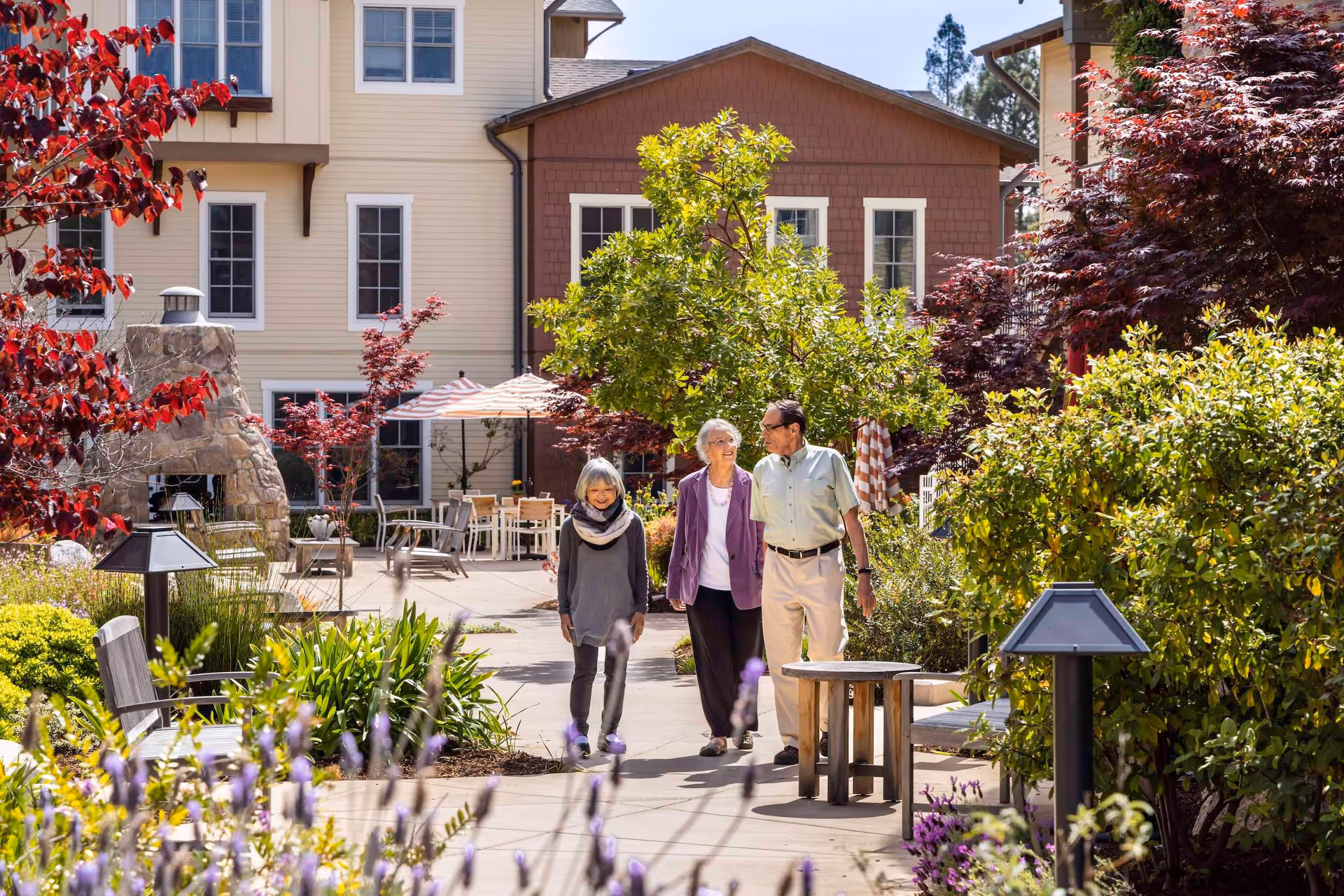 Three residents walking along a landscaped courtyard path with benches, flowering plants, and a multi-story building in the background.
