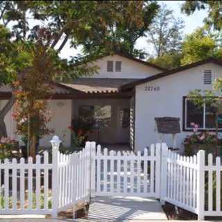 Front exterior view of a single-story residential building with a white picket fence, a pathway leading to the entrance, surrounded by trees and flowering plants.
