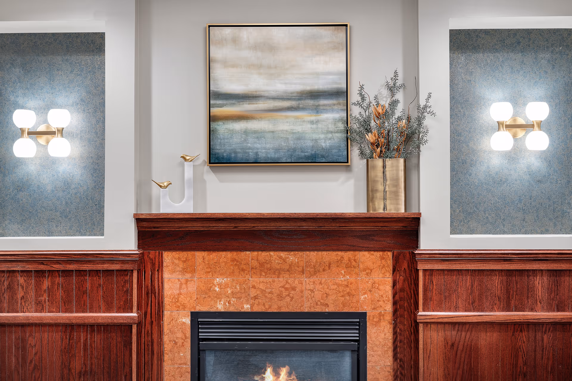 Wood-paneled fireplace mantel with a lit fireplace, abstract painting above, a brass vase with foliage, and glowing wall sconces.