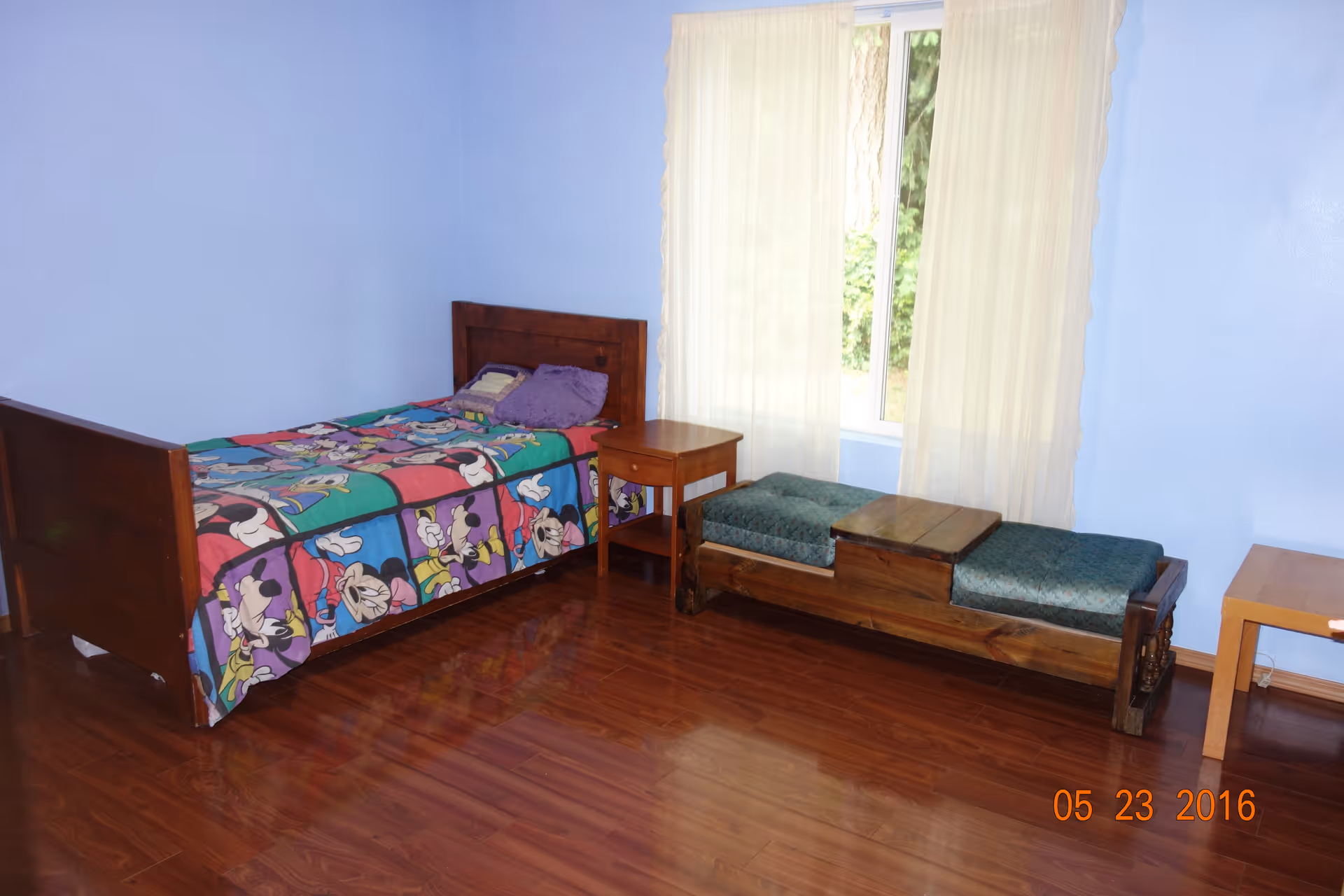 A bedroom with light blue walls and wooden flooring. There is a single wooden bed with a colorful cartoon-themed bedspread featuring Disney characters, a purple pillow, and a small wooden nightstand next to the bed. A wooden bench with green cushions and a small wooden table are placed near a window with sheer cream-colored curtains.