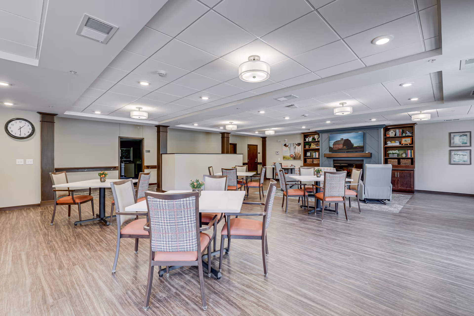 A spacious and well-lit common area with multiple tables and chairs arranged for dining or socializing. The room features wood flooring, a wall clock, ceiling lights, and a fireplace with built-in shelves displaying books and decorative items. There are also a couple of framed pictures on the wall and a comfortable armchair near the fireplace.