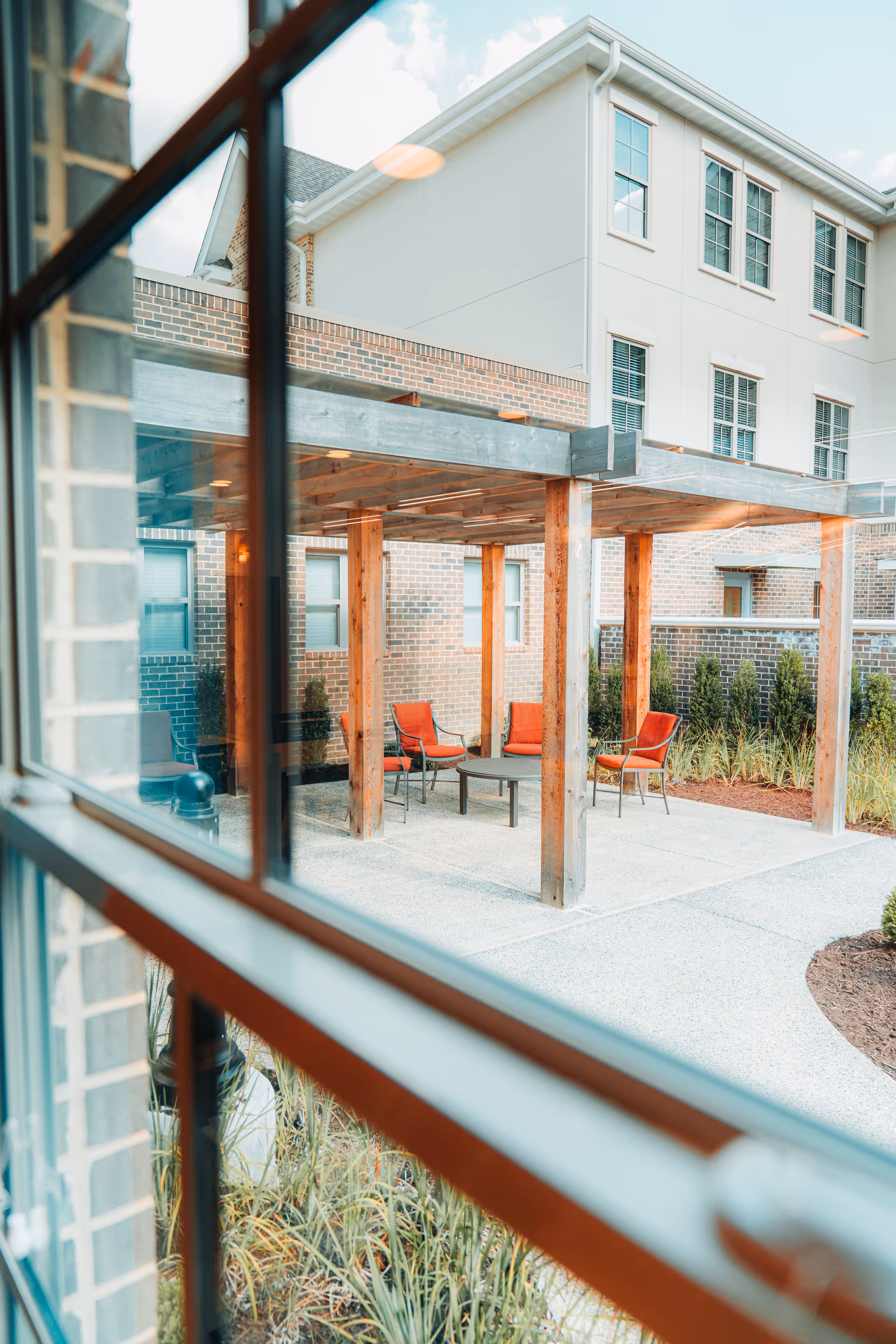 View through a window of an outdoor patio area with a wooden pergola, four orange cushioned chairs, and a round table. The patio is surrounded by a brick building and landscaping with plants and shrubs.