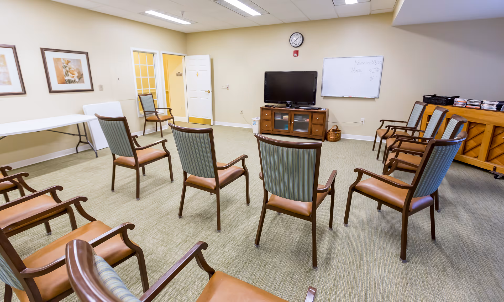 A room with chairs arranged in a semi-circle facing a television on a wooden cabinet. The room has beige walls, a clock above the TV, a whiteboard with writing, two framed pictures on one wall, and a piano with stacked items on top. There is a folding table and an open door leading to another room.