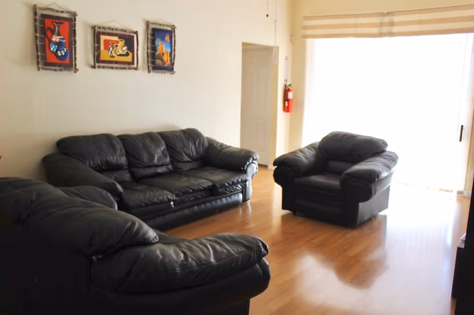 A living room with three black leather sofas arranged on a wooden floor. The room has light-colored walls with three framed artworks hanging above one sofa. There is a door and a large window with blinds letting in natural light.