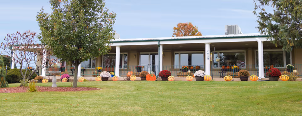 Front view of a single-story building with a covered porch decorated with pumpkins and potted mums and a grassy lawn with trees in front.