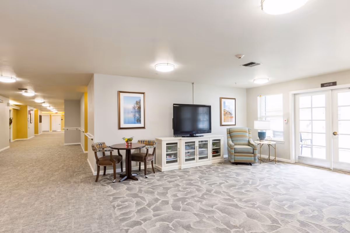 A bright and spacious common area in a senior living facility featuring a patterned carpet, a small round table with two chairs, a white TV stand with a flat-screen television, a striped armchair next to a side table with a lamp, framed artwork on the walls, and large windows with glass doors letting in natural light. A hallway with yellow and white walls extends to the left.
