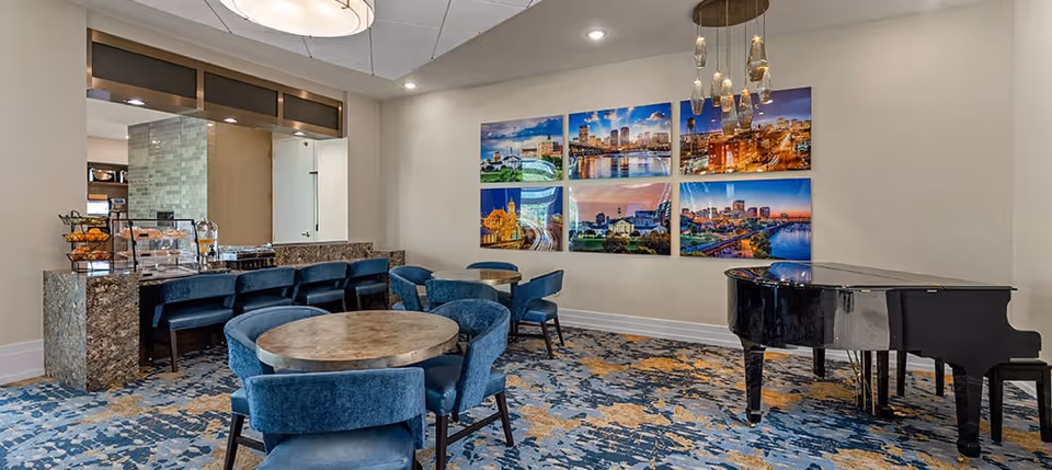 Communal dining/lounge area with round tables and blue chairs, a black grand piano, a buffet counter, and cityscape artwork on the wall.