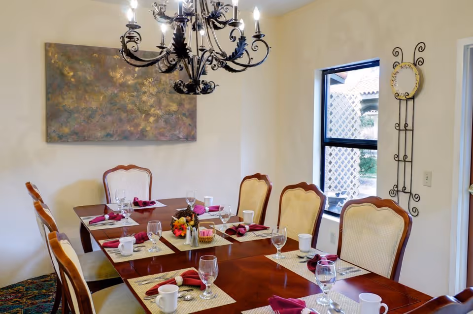 A dining room with a wooden table set for eight people. Each place setting includes a placemat, a white cup, a wine glass, silverware, and a folded burgundy napkin. A decorative centerpiece with flowers and pinecones is on the table. The room has cream-colored walls, a window showing an outdoor lattice, a large abstract painting on the wall, and a black ornate chandelier hanging above the table.