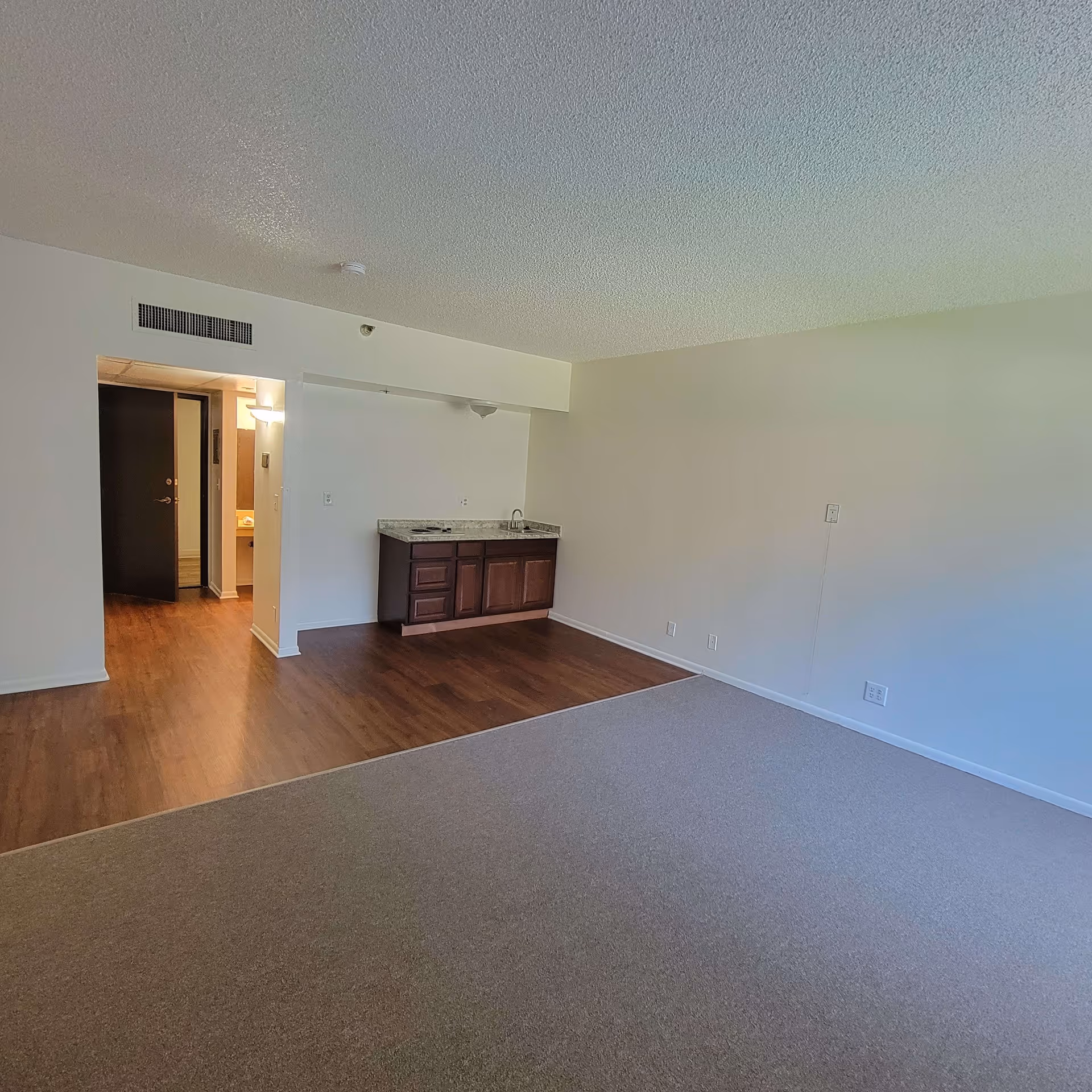 An empty interior room with beige carpet and wood flooring near a small kitchenette with dark wood cabinets and a granite countertop. There is an open doorway leading to a hallway and a bathroom with a sink and mirror visible. The walls are painted white and the ceiling has a textured finish.