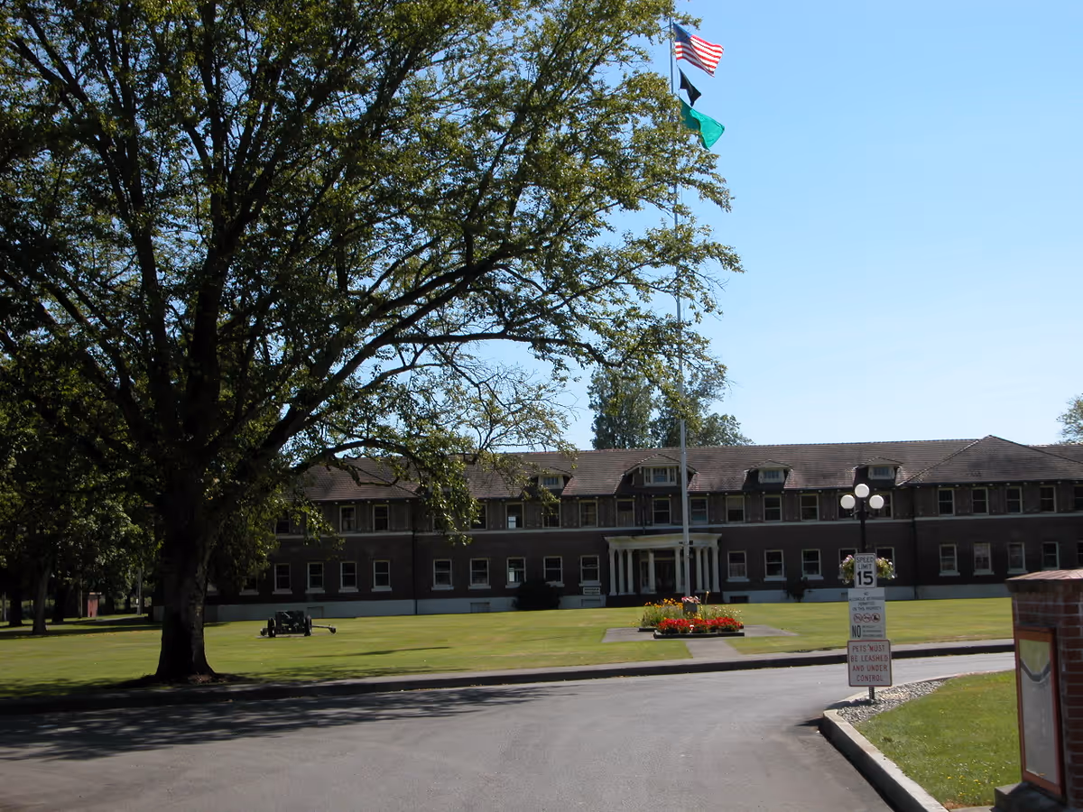 Exterior view of a large two-story brick building with multiple windows and a central entrance with columns. In front of the building is a well-maintained lawn with a flower bed and a tall flagpole flying three flags. A large tree is on the left side of the image, and a paved driveway curves in front of the building under a clear blue sky.