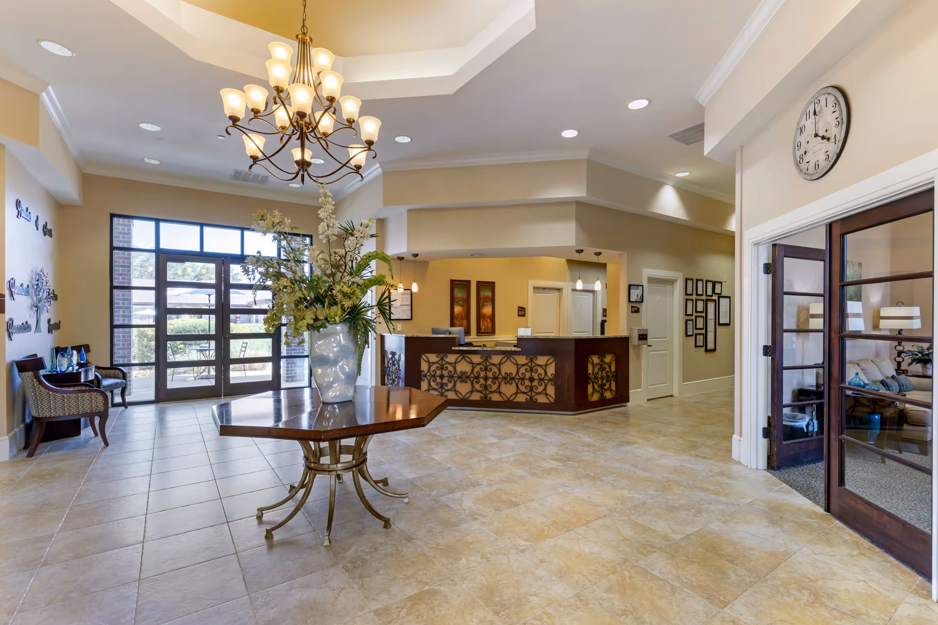 Spacious and well-lit assisted living facility lobby with a large chandelier hanging from a tray ceiling. A wooden table with a large vase of flowers is centered on the tiled floor. To the right is a reception desk with decorative metalwork and pendant lights overhead. Glass double doors lead to a cozy sitting area with a sofa and lamp. Large windows and a glass door provide natural light and a view of the outdoor area.