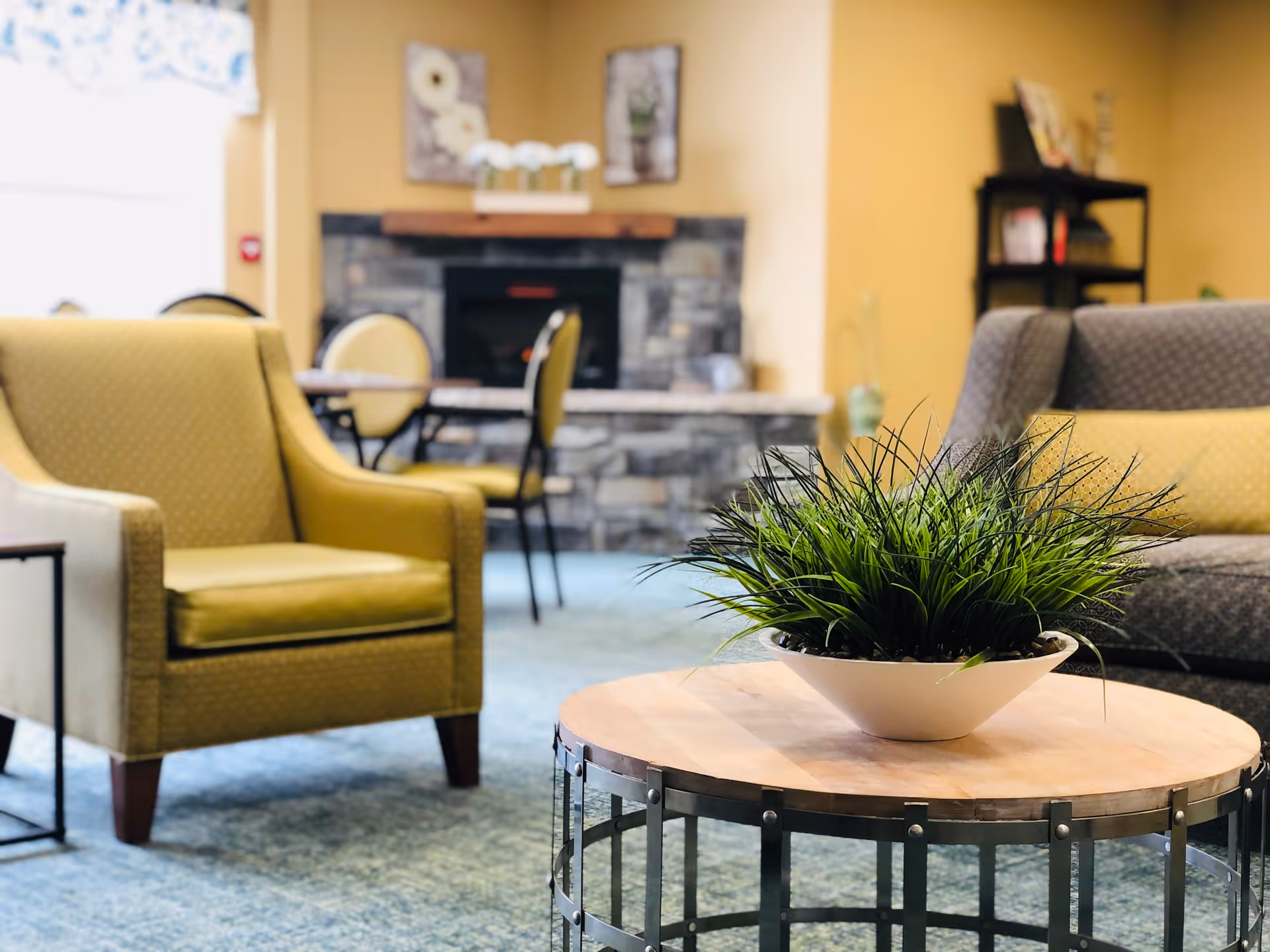 A cozy living room area with a round wooden coffee table featuring a white bowl filled with green plants. Surrounding the table are a yellow armchair and a gray sofa with a yellow cushion. In the background, there is a stone fireplace with decorative items on the mantel and a few chairs and tables near a window with a floral curtain.