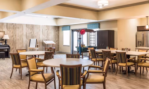A spacious dining room with multiple round and rectangular tables surrounded by wooden chairs with yellow cushions. The room features a light wood floor, a piano in the corner, and a cabinet near a doorway with windows. The walls are painted in neutral tones with a wood accent wall and a ceiling light fixture.