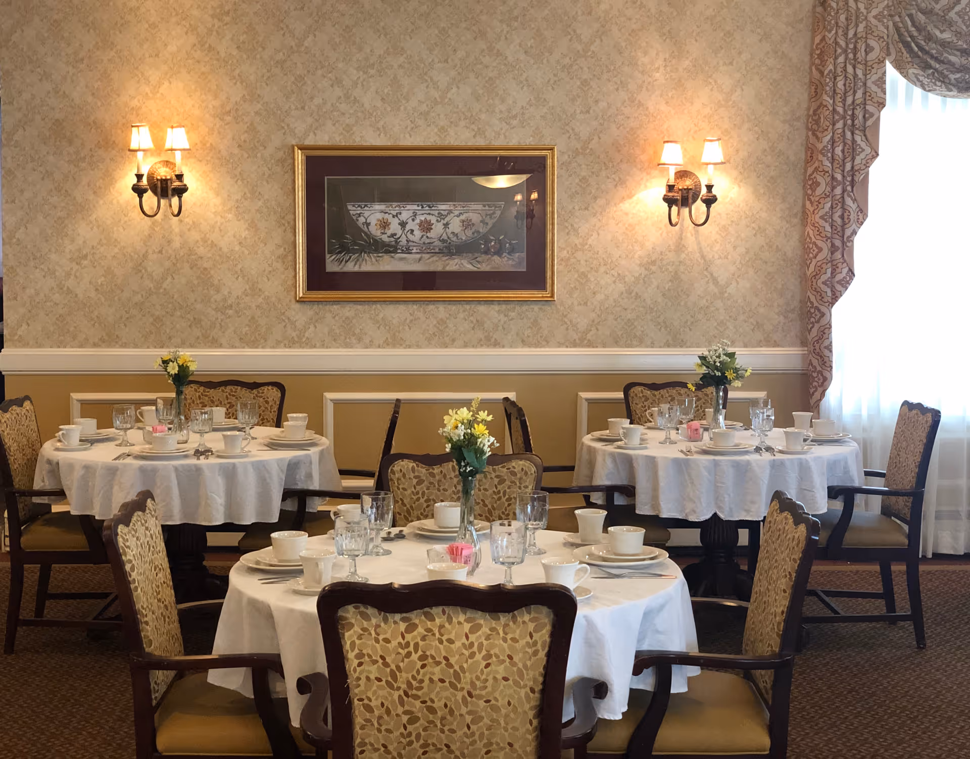 A dining room with three round tables covered with white tablecloths, each set with cups, plates, glasses, and silverware. Each table has a small vase with yellow flowers. The room has patterned wallpaper, two wall sconces with lampshades, a framed picture on the wall, and a window with patterned curtains allowing natural light.