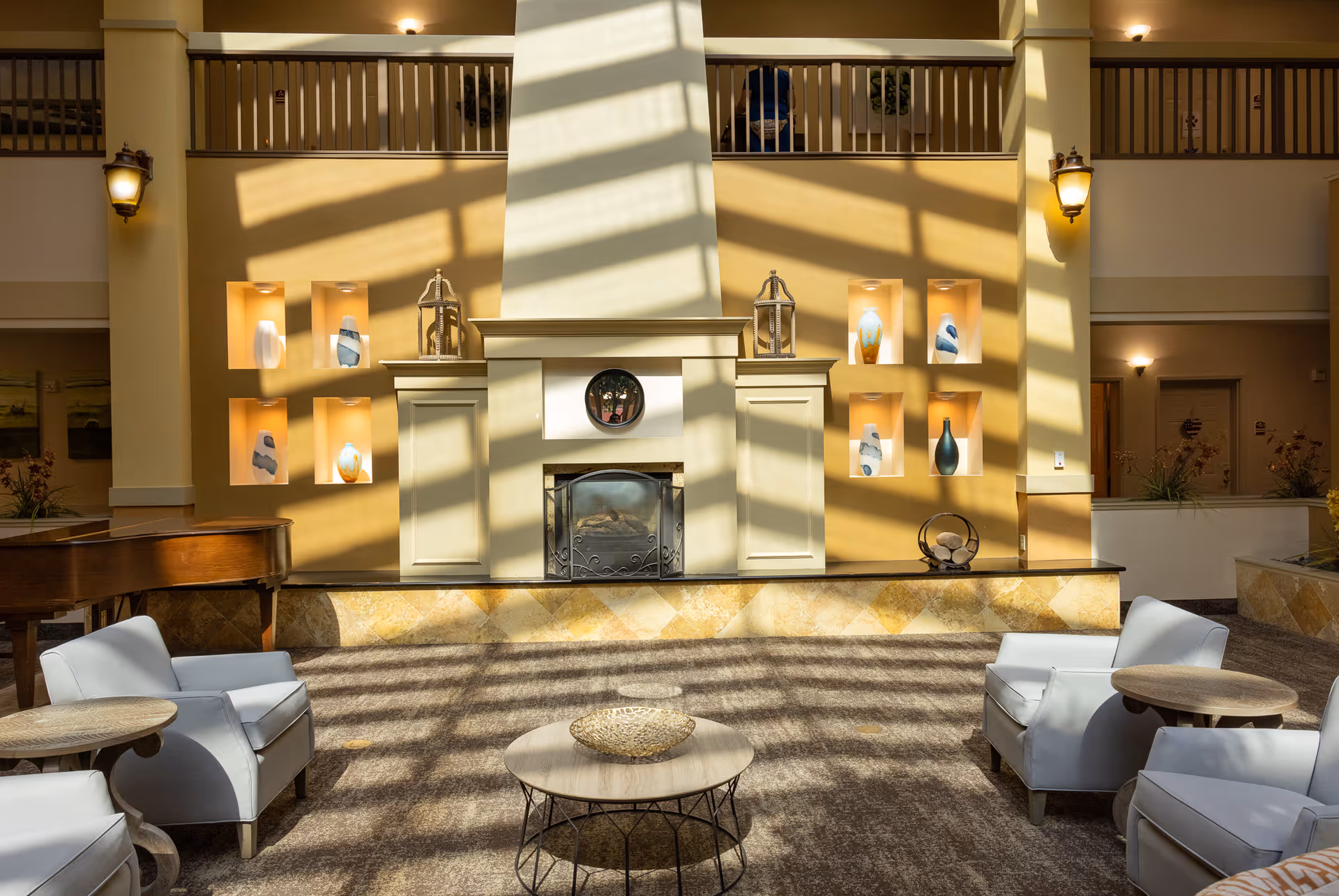 A bright and spacious senior living facility common area with a large fireplace centered on a beige wall featuring decorative niches with vases and sculptures. The room has four white armchairs arranged around two round wooden tables and a coffee table with a decorative bowl. Sunlight streams through windows, casting shadows on the carpeted floor. A grand piano is partially visible on the left side.