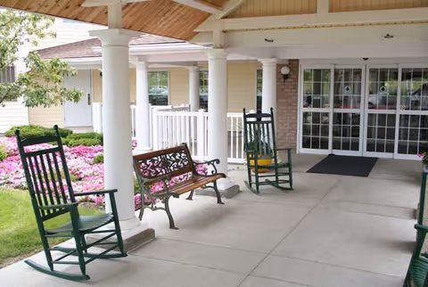Covered outdoor patio area with white columns, two green rocking chairs, a wooden bench with decorative metal armrests, and a garden with pink flowers in the background. The entrance to a building with large glass doors is visible.