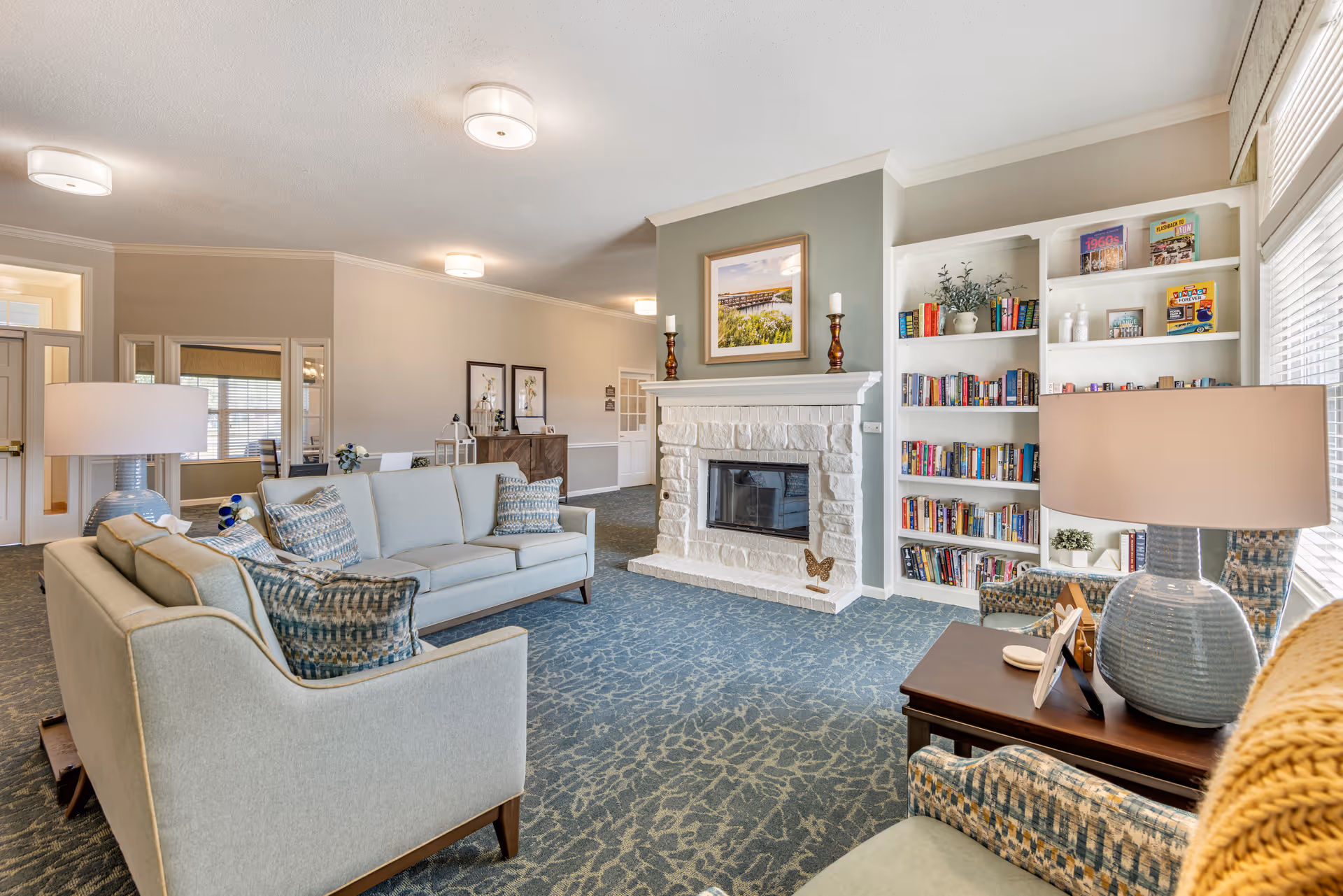 A cozy living room area in a senior living facility featuring two light blue sofas with patterned cushions, a white stone fireplace with a framed picture and candles on the mantel, a built-in bookshelf filled with books and decorative items, a side table with a blue lamp, and large windows allowing natural light to fill the space.
