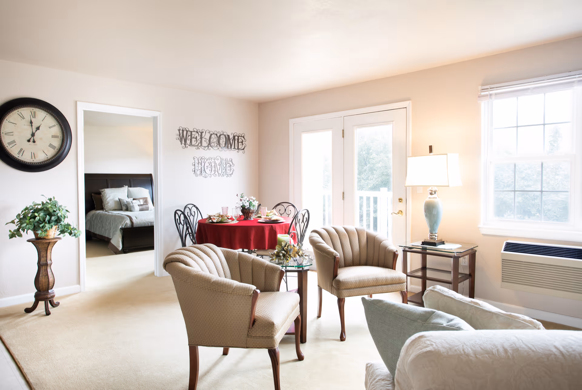 Sunlit living room with upholstered armchairs, a small round dining table with a red tablecloth, and a doorway to a bedroom.